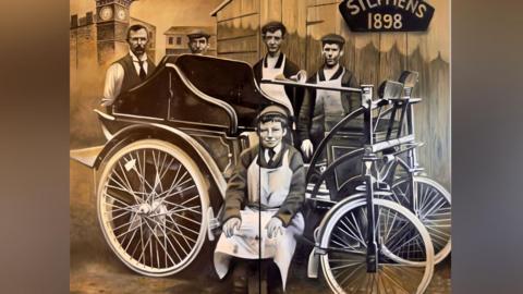 A painting showing four men wearing Victorian clothes and aprons standing around a primitive Victorian motor car, with the words "Stephens 1898" painted on the mural