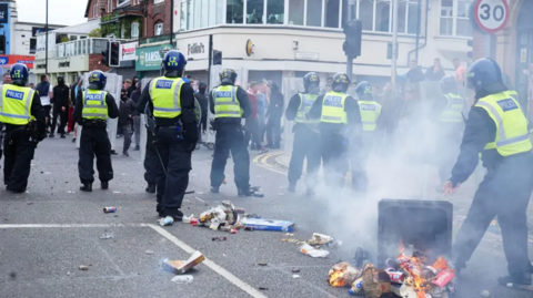 A line of police in riot gear facing a group of people. A bin has been tipped over and is on fire and rubbish is all over the road.