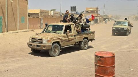 FLA fighters sitting in a pickup truck on a sandy road, with another in the distance behind them. to the left are buildings lining the road. In the front right, there is an orange metal barrel
