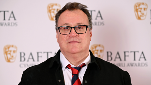 Russell T Davies attending the 2024 BAFTA Cymru awards at the International Convention Centre Wales. Behind him is a sign that says BAFTA Cymru Awards. He is wearing a white shirt, black jacket and a red and dark blue tie. Davies has short brown hair and wears glasses.