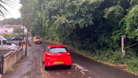 A car and a van parked on a residential street, in grey rainy weather. There are houses on the left, and dense green woodland on the right. A wooden footpath sign can be seen leading into the woods. 