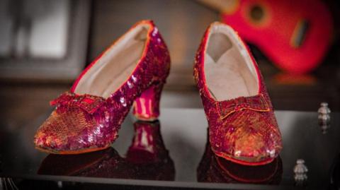 A close-up of the ruby red sequin shoes worn by Judy Garland. They feature dark red sequins sewn in chevron strips across the front of the shoes, with red bows sewn on and thick block heels at the back, with a light coloured insole