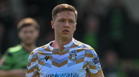 Matt Curley is wearing a white Tamworth FC away shirt and is about to pass the ball. The ball is out of shot. Curley is also wearing a white bandage on his left wrist. There is an opposition player in green in the background but the image is blurred.