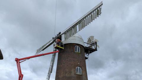 Under a dark grey sky, the top of the windmill, which is a round red brick building with a white dome on top, has a cherry picker lifting men to the white sails as they remove one