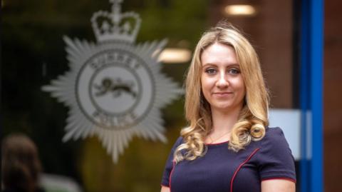Ellie Vesey-Thompson, a young woman with long blonde hair and wearing a blue dress with red piping, stands in front of a glass door with the logo of Surrey Police on it.