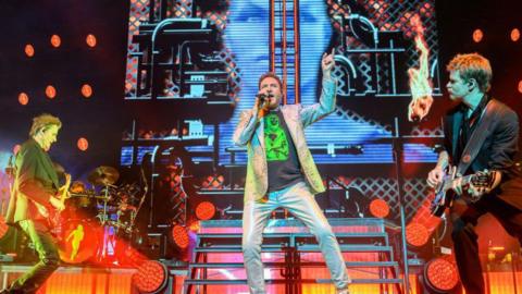 Duran Duran are pictured performing on stage. The lead singer Simon Le Bon holds a microphone to his mouth as he singers while two guitarists stand on each side of him playing.