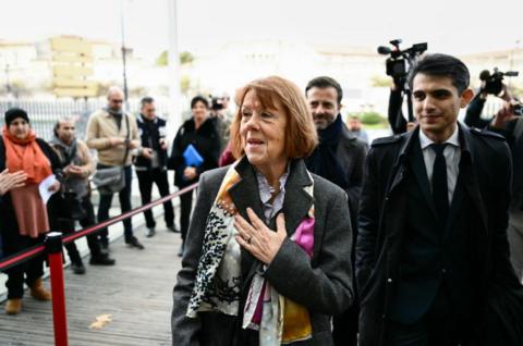 Gisèle Pelicot wears a grey coat and a colourful scarf and smiles at people standing near her with a hand on her heart