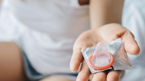 A woman is holding a condom in the palm of her hand. Her hand and the condom is in focus and in the background you can see the woman wearing a white top and blue shorts.