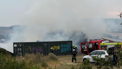 A picture of the scene where the fire took place. There is a large metal container in front of the smoke which is rising up. There is firefighters pictured tackling the blaze in distance.
