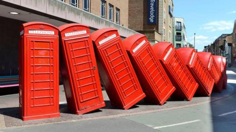 A outdoor sculpture on a suburban street consisting of a row of 12 red phone boxes that have collapsed onto each other.