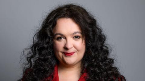 Ashley Storrie, a woman with long dark hair, wearing a red jacket. She is staring into the camera