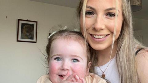 Sian Kennedy, who has long blonde hair, holding her little girl Sienna. Both are smiling at the camera.