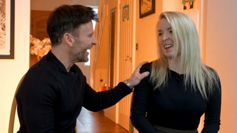 A man and woman gaze into each others eyes smiling, the man has his hand on the woman's shoulder