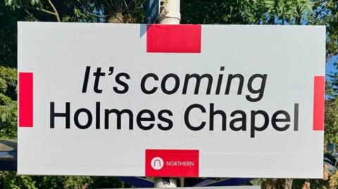 Railway station sign reading: 'It's coming Holmes Chapel'.