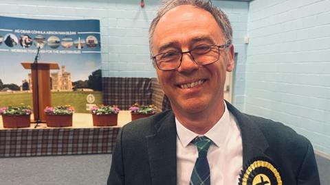 Alasdair Allan is pictured indoors. He has grey hair and is wearing glasses. He has a dark suit jacket over a white shirt and a tartan tie. He is wearing a yellow and black rosette.