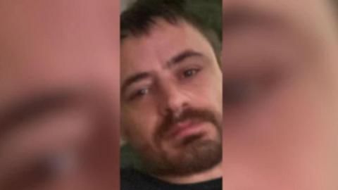 A close up image of a man's face. He has short brown hair and a brown beard. There are blurred borders on either side of the image. 