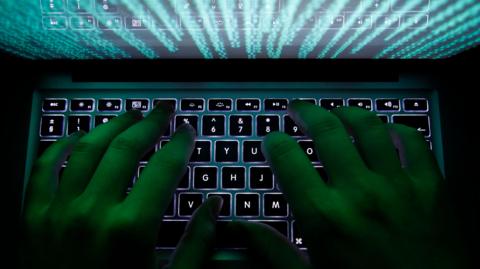A man types on a keyboard with green code illuminating his fingers green.