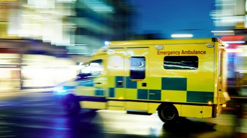 Yellow and green ambulance speeding through night with 'Emergency Ambulance' written on the side.