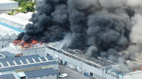 Drone shot showing bright orange flames coming through the roof of the factory, and thick black smoke belching into the sky.