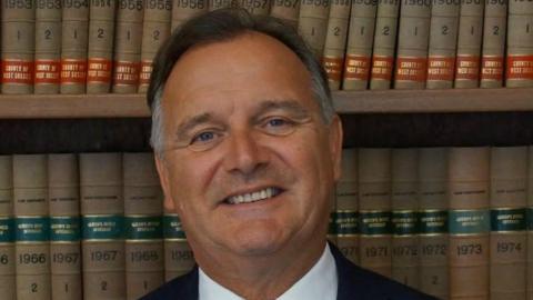 Paul Marshall has short brown hair and wears a black suit with a white shirt and red spotted tie.