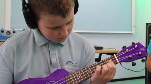 A boy in a light blue polo wears black headphones and is playing a purple guitar. 