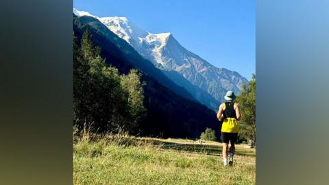 A man runs along a green field in the sun with picturesque mountains in front 