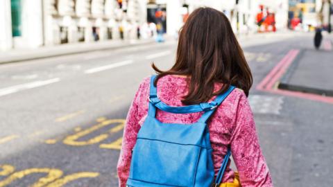A young woman with brown hair, a pink top and a blue packpack walks down a street