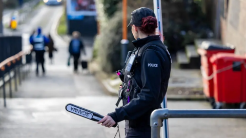 A female police officer stands in a street holding body scanning equipment. The officer is in black uniform with her hair tied back and a cap on. She is looking away form the camera down the street. She is stood near some recd bins and people can be seen further on down the street.
