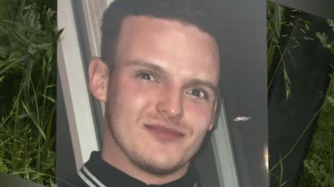 A large print out of a photo of Stephen Morrisson is propped up against foliage. He is wearing a black polo with white stripes on the collar. He has a slight smile and has dark brown hair.