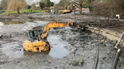 A yellow digger is submerged in mud and silt. There is another digger in the background, trees and the top of a house.