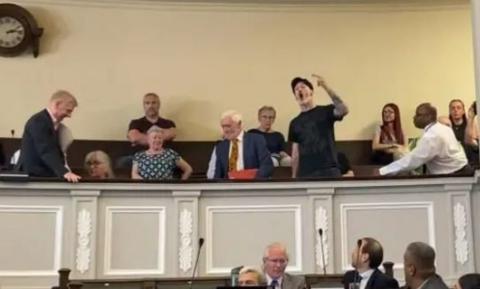 A video still of members of the public sat in a gallery above councillors in the city council chamber. One man, dressed in a black T-shirt and a black hat, is stood up and gesturing with a raised arm towards the councillors.
