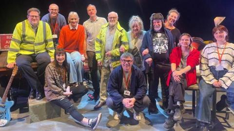 A group of people dressed in many different ways, from fluorescent jackets to casual attire, sitting on the set of a play on stage. On the left hand side is a Road Ahead Closed sign and a blue electric guitar.