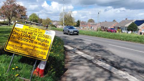 Yellow roadworks sign on a grass verge on the side of the road. There's a green verge and houses and bungalows in the background.