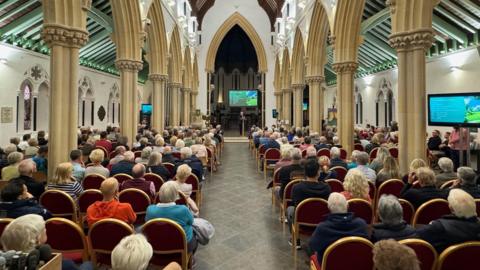 A church hall with people sat in rows looking at Helen Powell delivering a presentation about the campaign