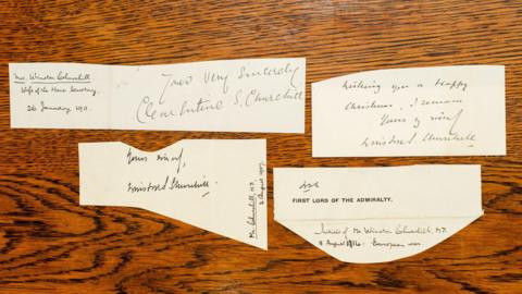 Four small pieces of paper that have been cut out sit on a dark wood table but not very much of the table is shown. The photo is taken from directly above. Each piece of paper contains handwritten signatures by Winston Churchill.