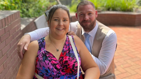 Sandy is wearing a pink dress and is sat down with her husband behind her, who is wearing a shirt, tie and waistcoat