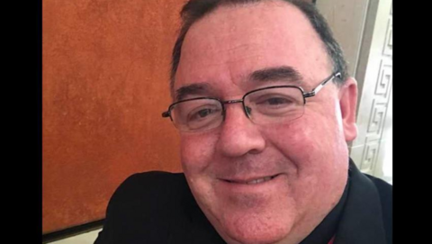 Shows a man wearing glasses and dark clothing smiling