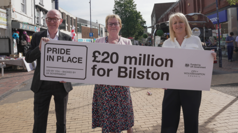 Pat McFadden MP, Josie Kelly and councillor Linda Leach stand in a street in Bilston town centre holding a board or banner saying £20m for Bilston, Pride In Place. 