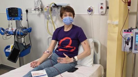 A woman with short dark hair and wearing a face mask sits on a hospital bed in blue T-shirt and jeans, hooked up to a red tube connected to a transfusion machine.