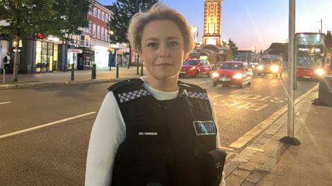 Surrey Police and Crime Commissioner Lisa Townsend wears a white top and a black police stab vest as she stands in Epsom town centre at dusk on a summer evening.