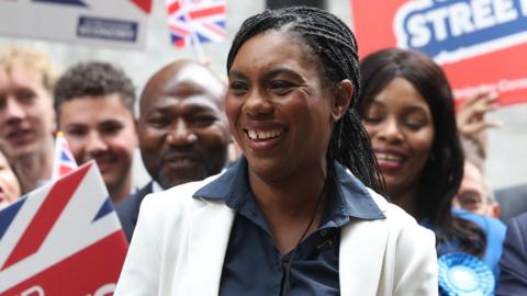 A grinning Kemi Badenoch celebrates a victory in Westminster with activists waving union jack banners and wearing blue Tory rosettes
