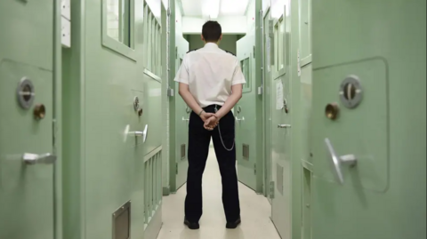 A male prison officer standing in a prison, in uniform, with his hands behind his back and his back to the camera. He's standing in a narrow corridor between two rows of cells with light green doors.