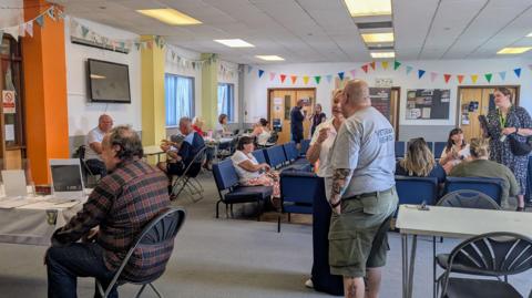 A picture of a room full of people. It is a large room with a number of seats and people stood around. 