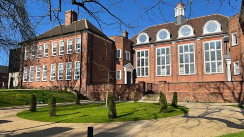 Large victorian listed building with orange bricks and rectangular victorian windows set behind a courtyard with green space 