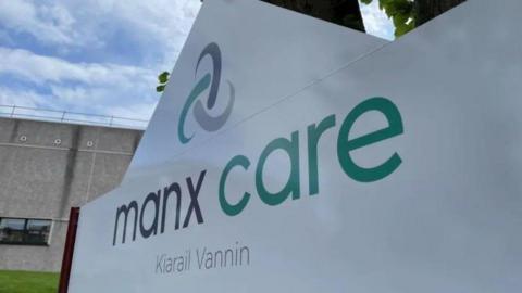 A large white Manx Care sign which has the organisation's name in English and Manx in green and grey lettering along with its logo.