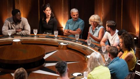 Contestants on The Traitors sitting behind a round table during an episode. One woman can be seen talking as the others listen to her. 