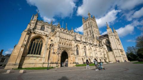 A picture of two people standing in front of Gloucester Cathedral. The sky is blue and there are some clouds in the sky.