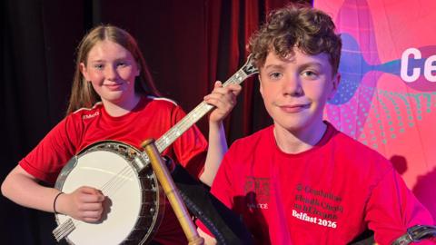A girl with long light brown hair and a boy with brown hair both holding musical instruments and wearing red t-shirts.