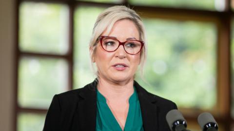 Michelle O'Neill in a black blazer and green blouse with red glasses. She has blonde hair tied up behind her head and is standing in front of two black microphones.