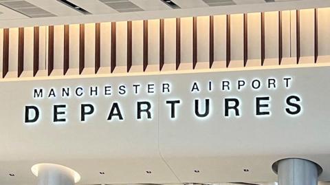 Sign for Manchester Airport Departures on a cream ceiling above the airport gate
